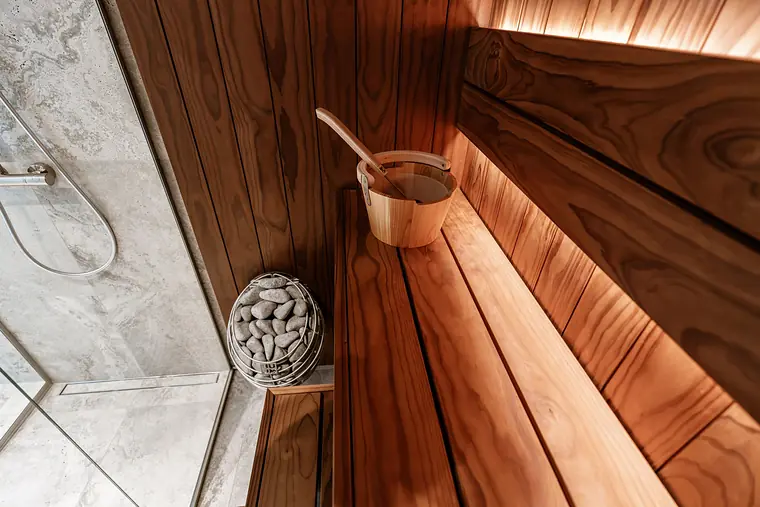 A warm timber sauna: slatted benches, a stone heater and a wooden bucket and ladle in the window light